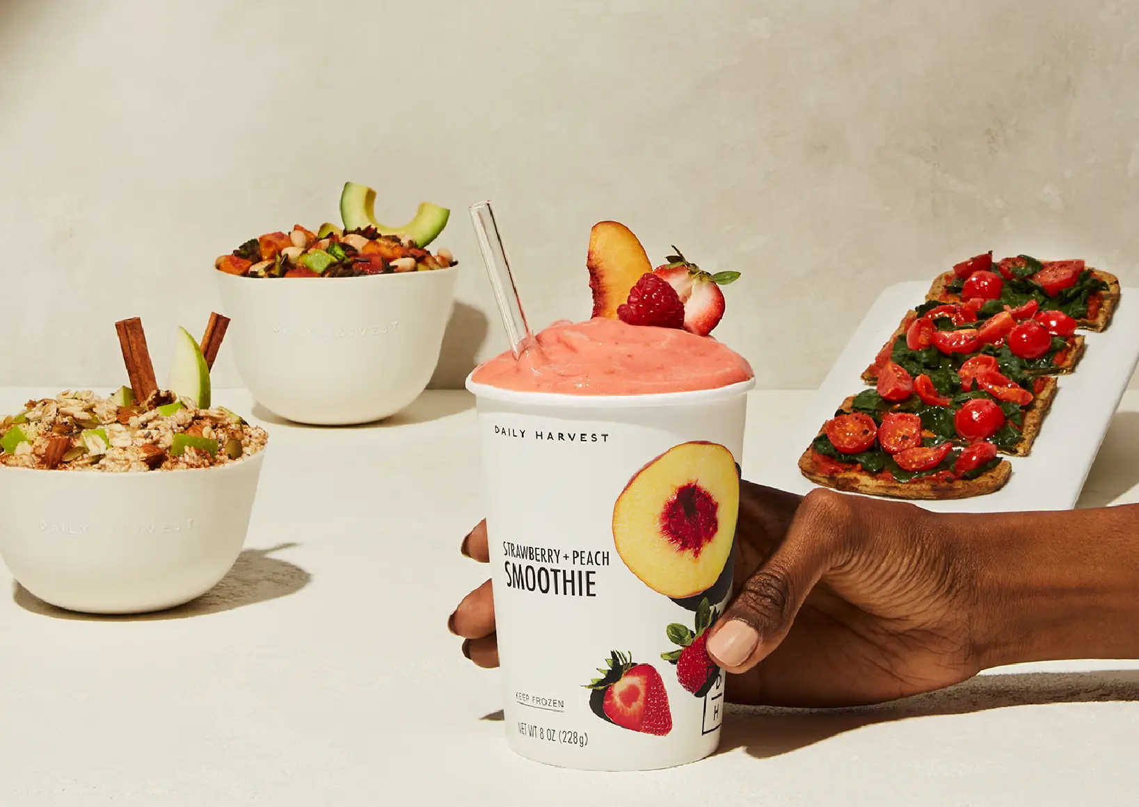 A person's hand holding a Daily Harvest smooth with two bowls and a flatbread behind it.