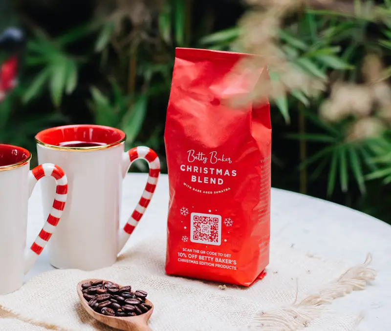 A tabletop decorated with two candy cane-rimmed mugs, a wooden spoon full of coffee beans, and a red package of coffee beans with a QR Code